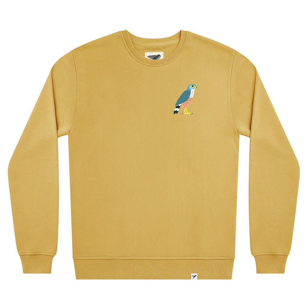 A mustard yellow crewneck sweatshirt featuring a small Merlin embroidered bird graphic on the left chest, displayed on a clean white background.